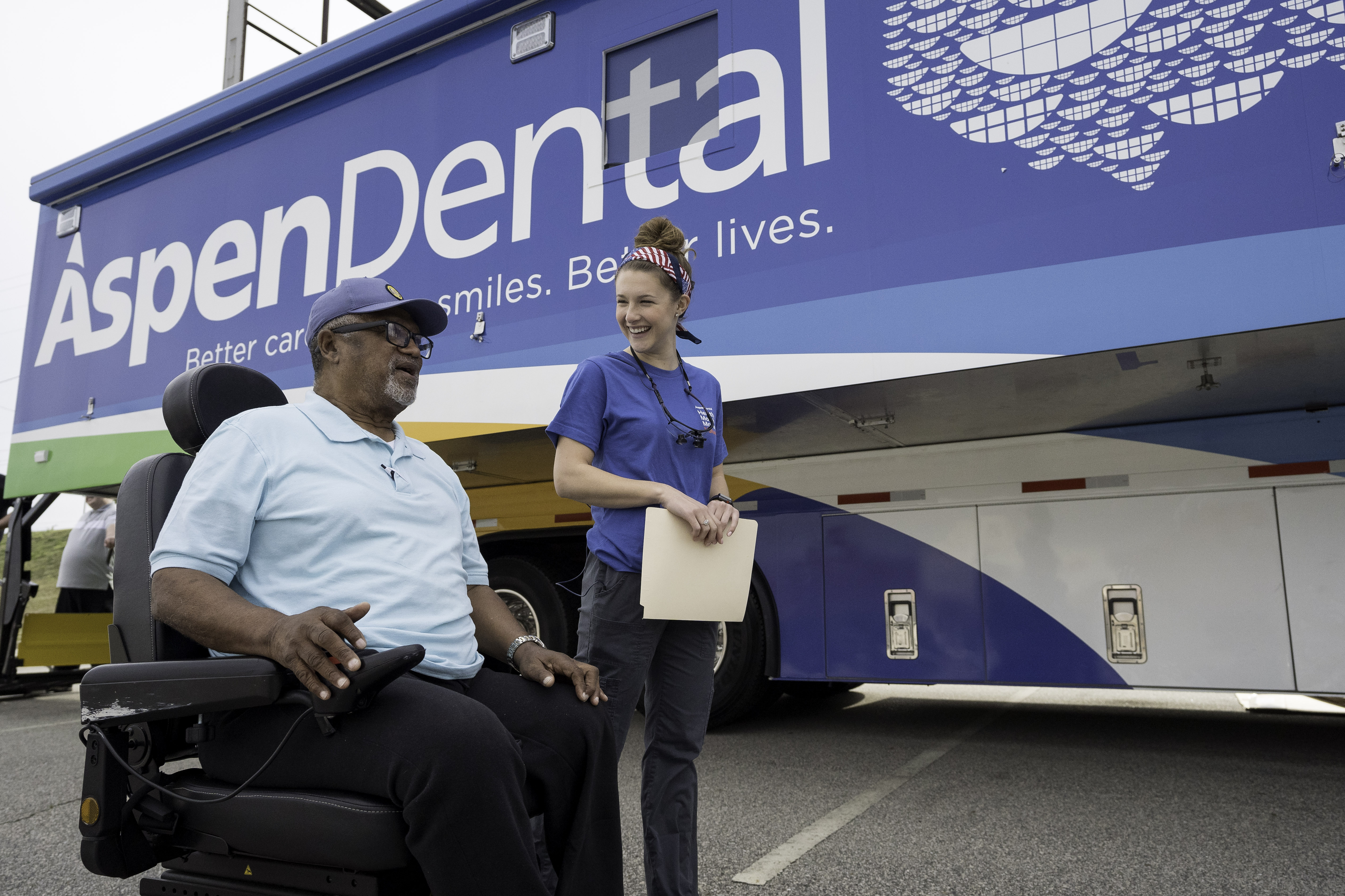 A man in a wheelchair next to a young female dental volunteer in front of the aspen dental mouth mobile bus